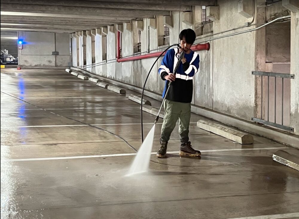 Crew member pressure washing a covered garage level, wet floor reflecting the lights