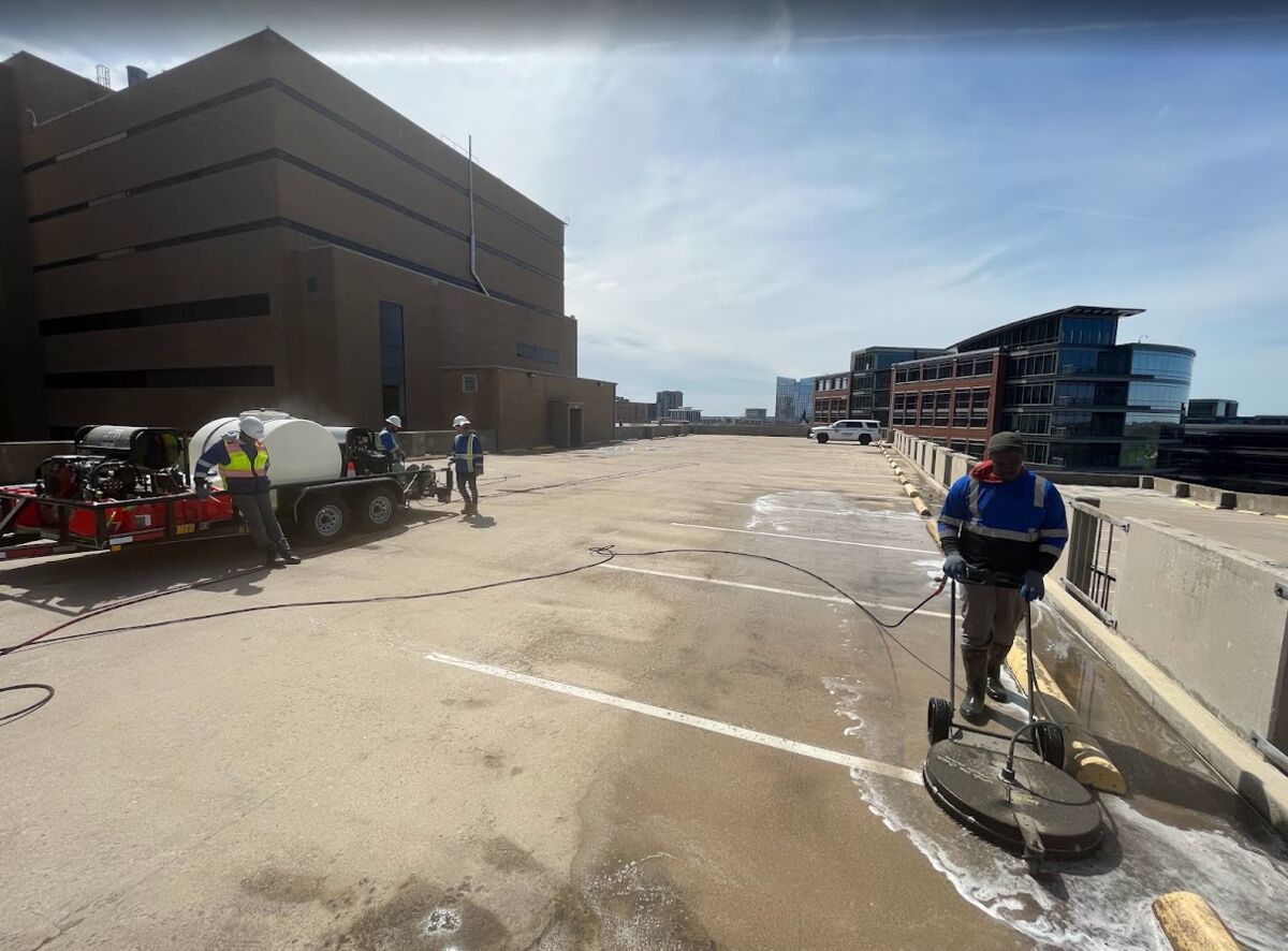 Four-person crew washing a rooftop parking deck with the trailer rig and downtown buildings behind