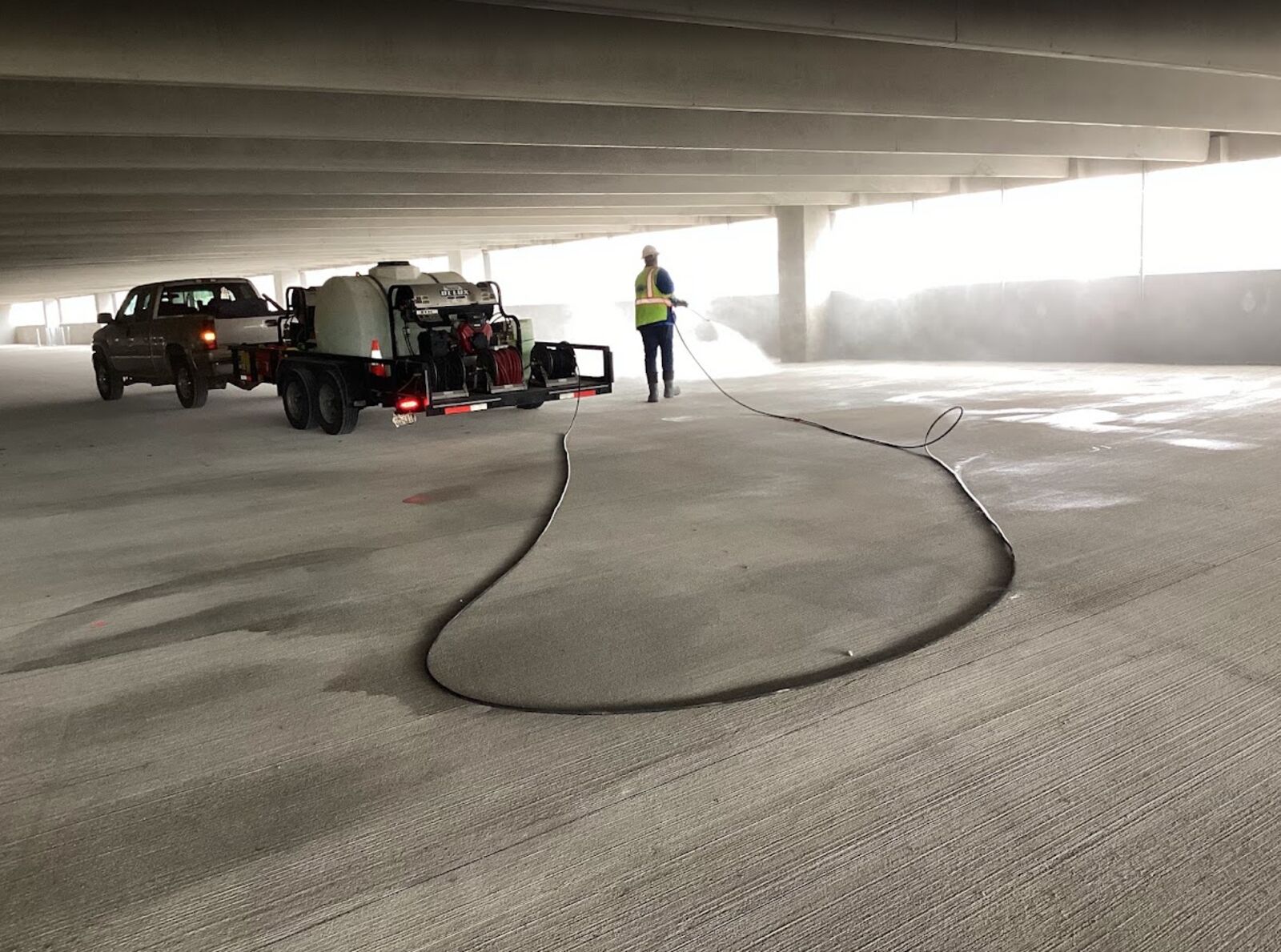 Nitty Gritty crew member pressure washing a covered parking garage level, with the hot-water trailer rig and pickup parked on the deck