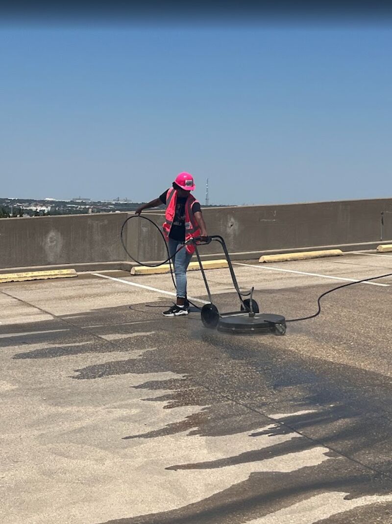 Crew member in a pink hard hat running a surface cleaner on a rooftop garage deck, leaving a clean path beside stained concrete